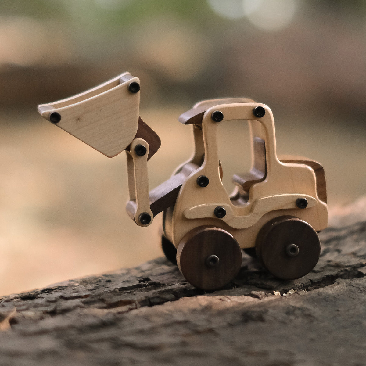 Maple Digger wooden toy on a weathered log in the woods, pale maple body and raised loader bucket with dark walnut wheels