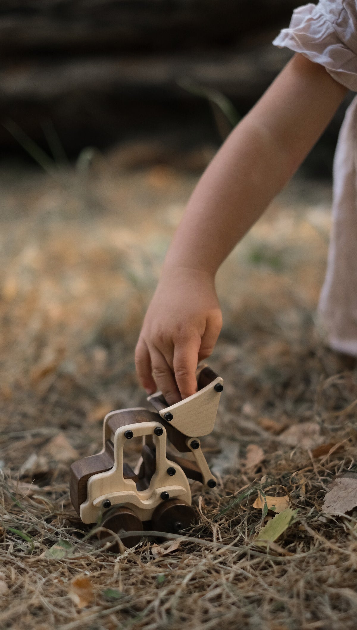 Child's hand lifting the maple bucket of the Maple Digger on dry grass and fallen leaves, walnut wheels and seat back visible