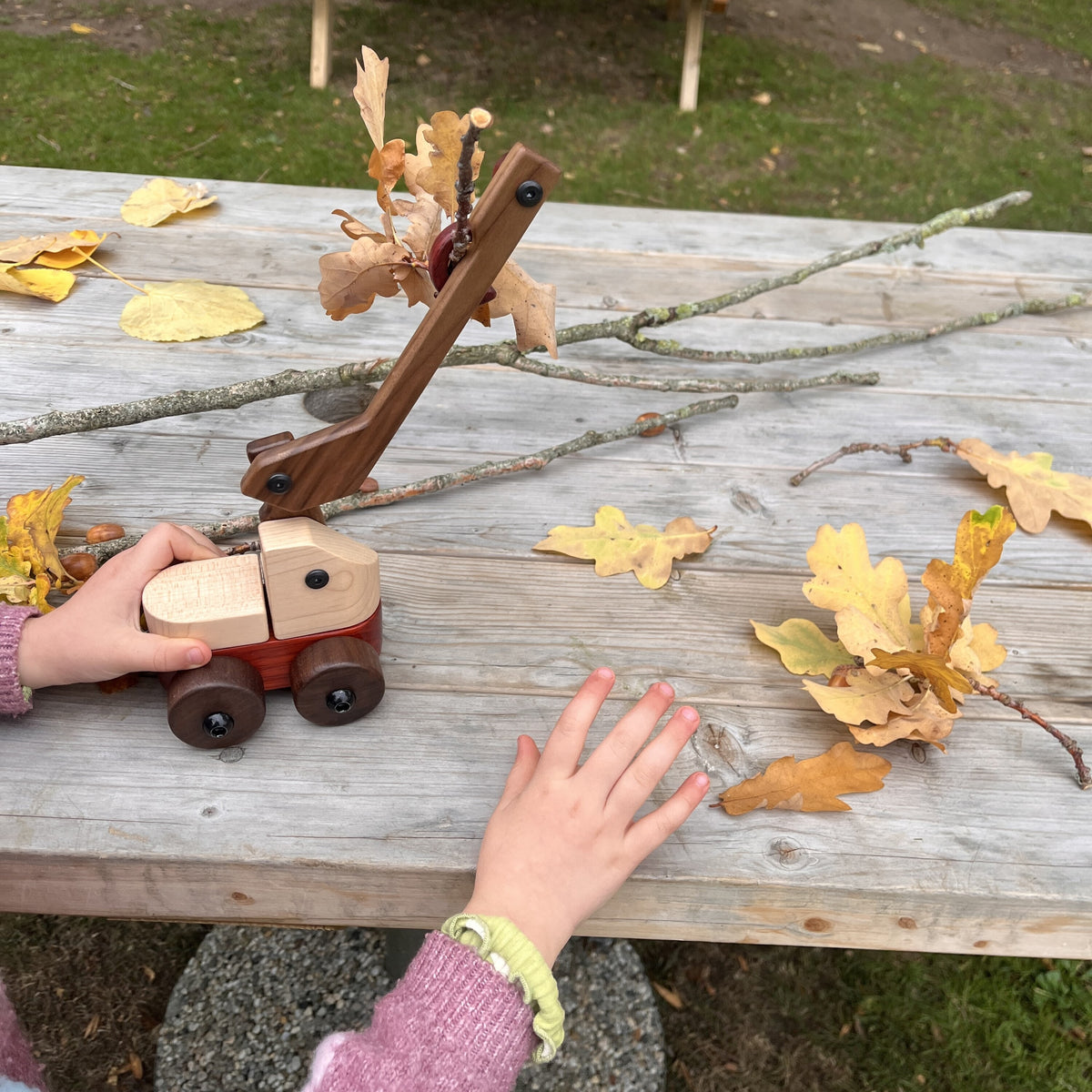 Child's hands playing with the Walnut & Maple Crane on an outdoor wooden table, autumn oak leaves caught on its hook, with twigs and fallen leaves