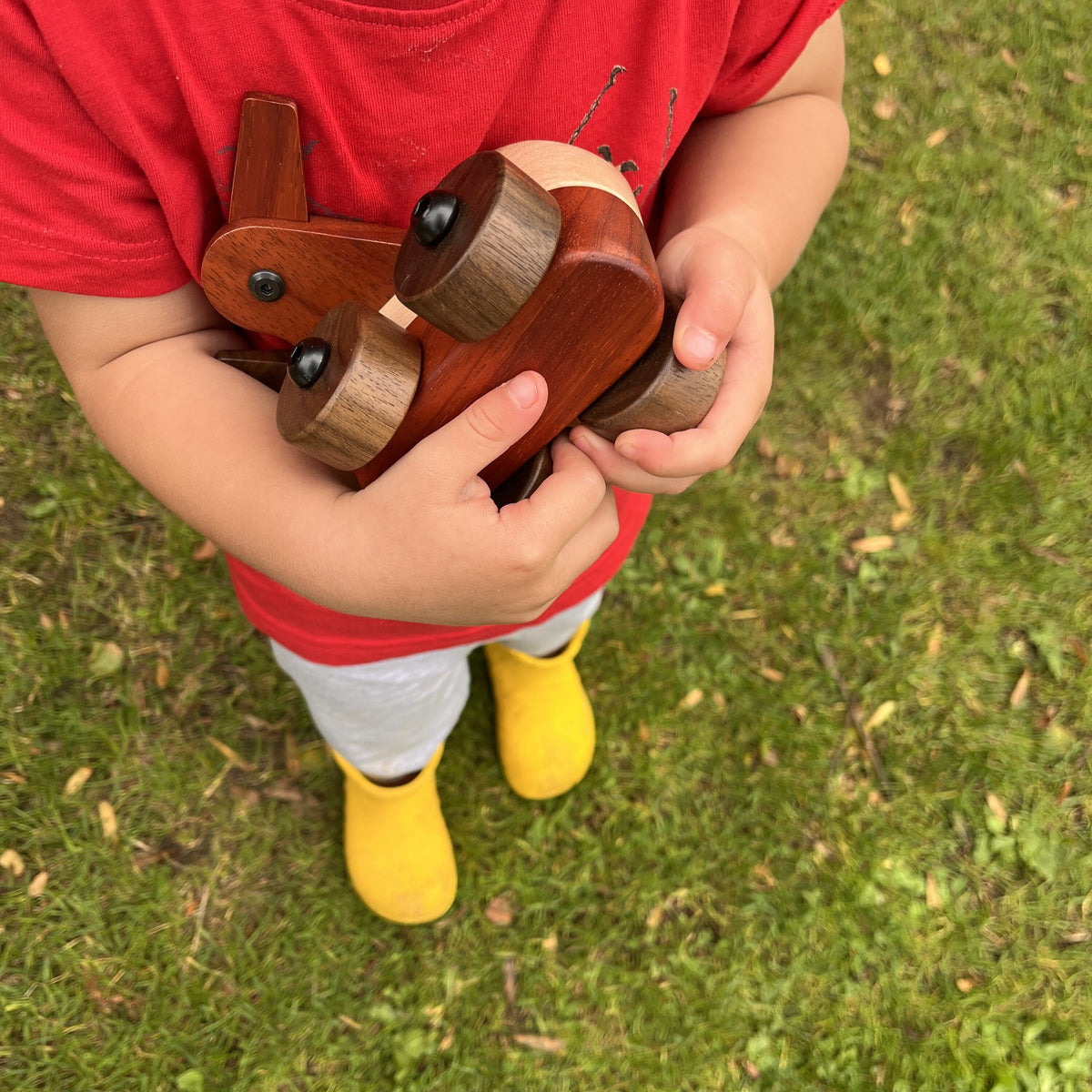 Child in a red T-shirt and yellow wellies holding the Walnut & Padauk Aeroplane against their chest, standing on grass