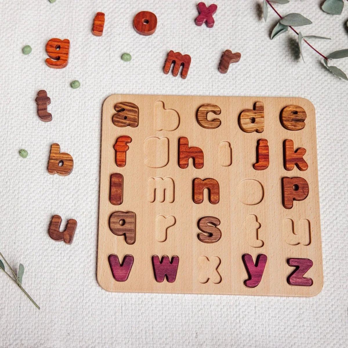 Oak & Sprout lowercase alphabet puzzle partially assembled, tigerwood and purpleheart letter pieces scattered on linen