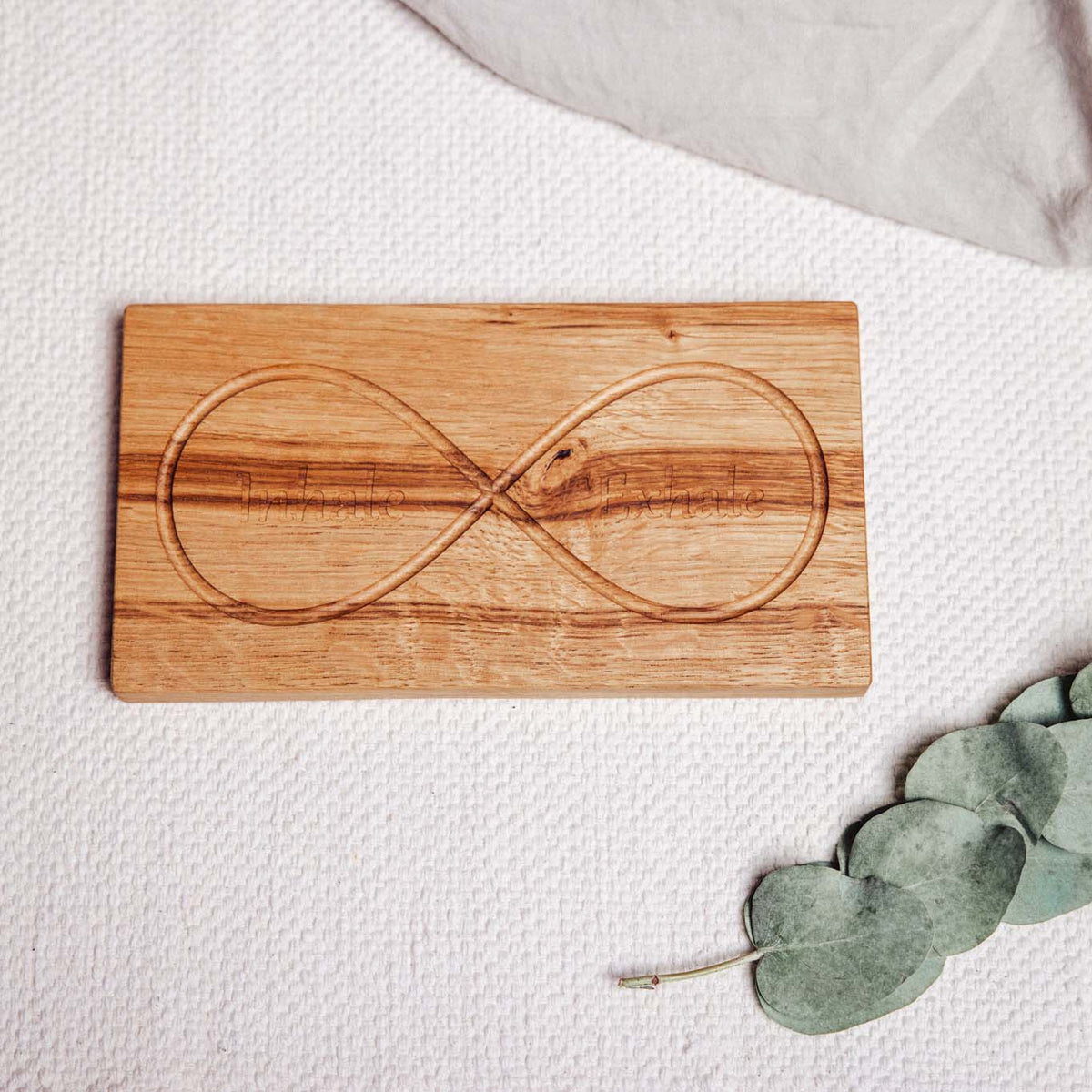Infinity Breathing Board in oak, flat-lay with engraved inhale and exhale loops visible, styled with eucalyptus on cream knit