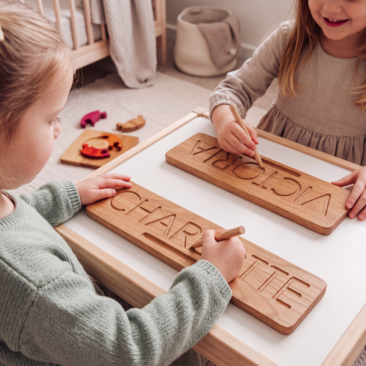 Two young sisters at a play table each tracing their own name, CHARLOTTE and VICTORIA, on solid oak name tracing boards with short oak styluses