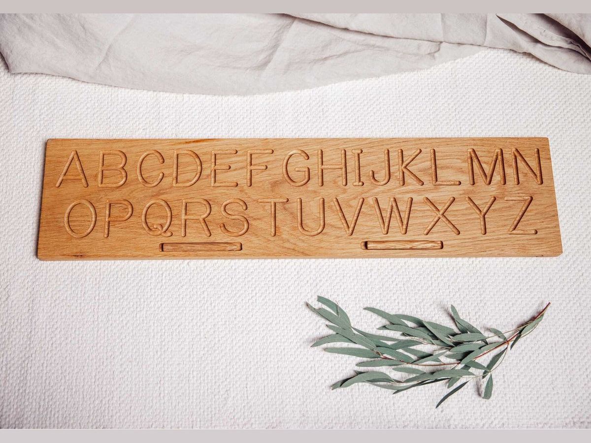 Solid oak alphabet tracing board with the capital letters A to Z routed in two rows and two stylus grooves, laid on white woven cotton beside a sprig of eucalyptus
