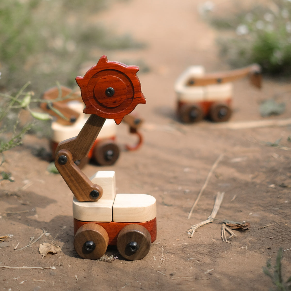 Padauk & Maple Trench Digger on a dirt path with its padauk cutter wheel raised, two more Oak & Sprout wooden vehicles blurred behind