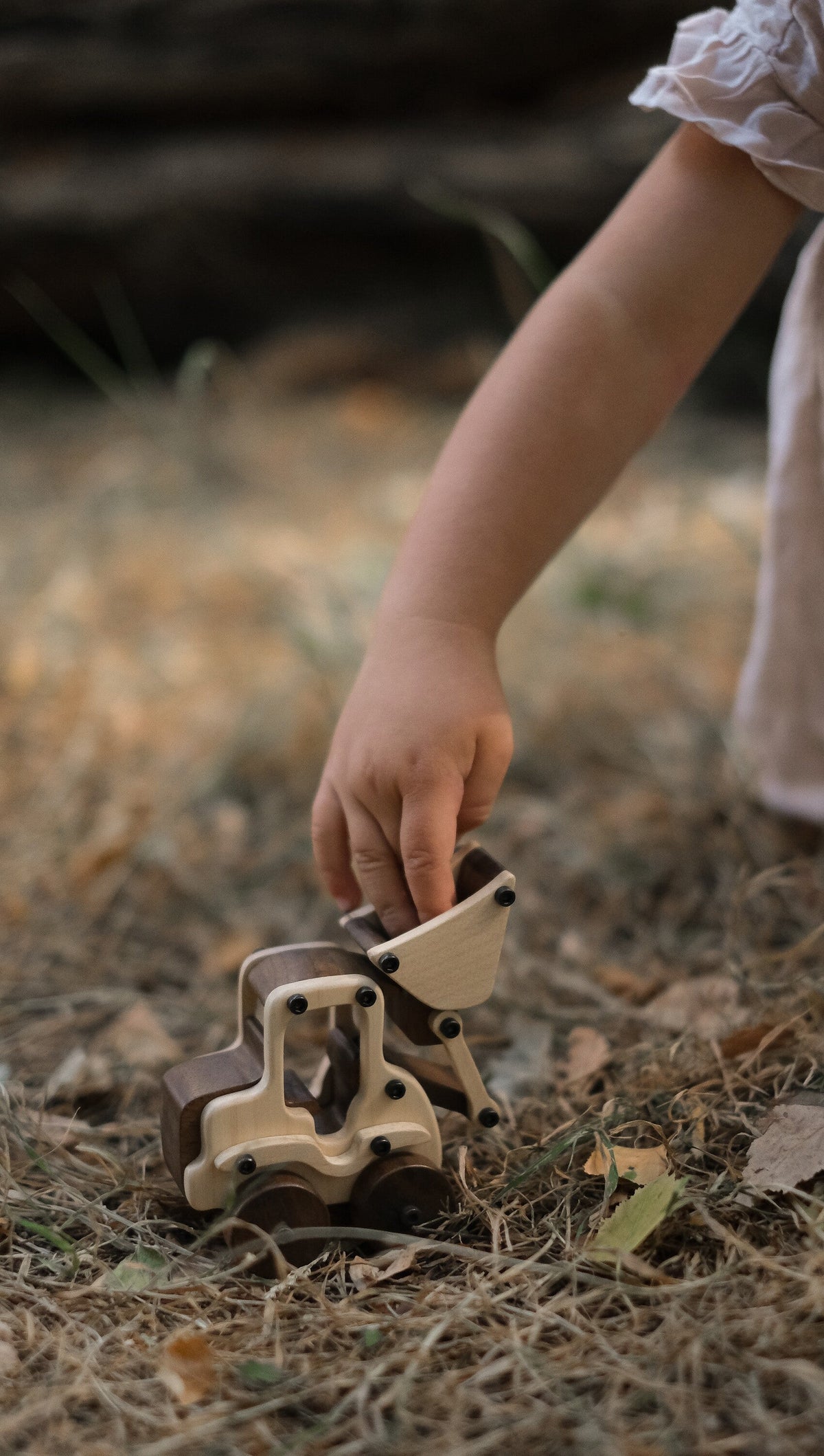 Child's hand lifting the maple bucket of the Maple Digger on dry grass and fallen leaves, walnut wheels and seat back visible