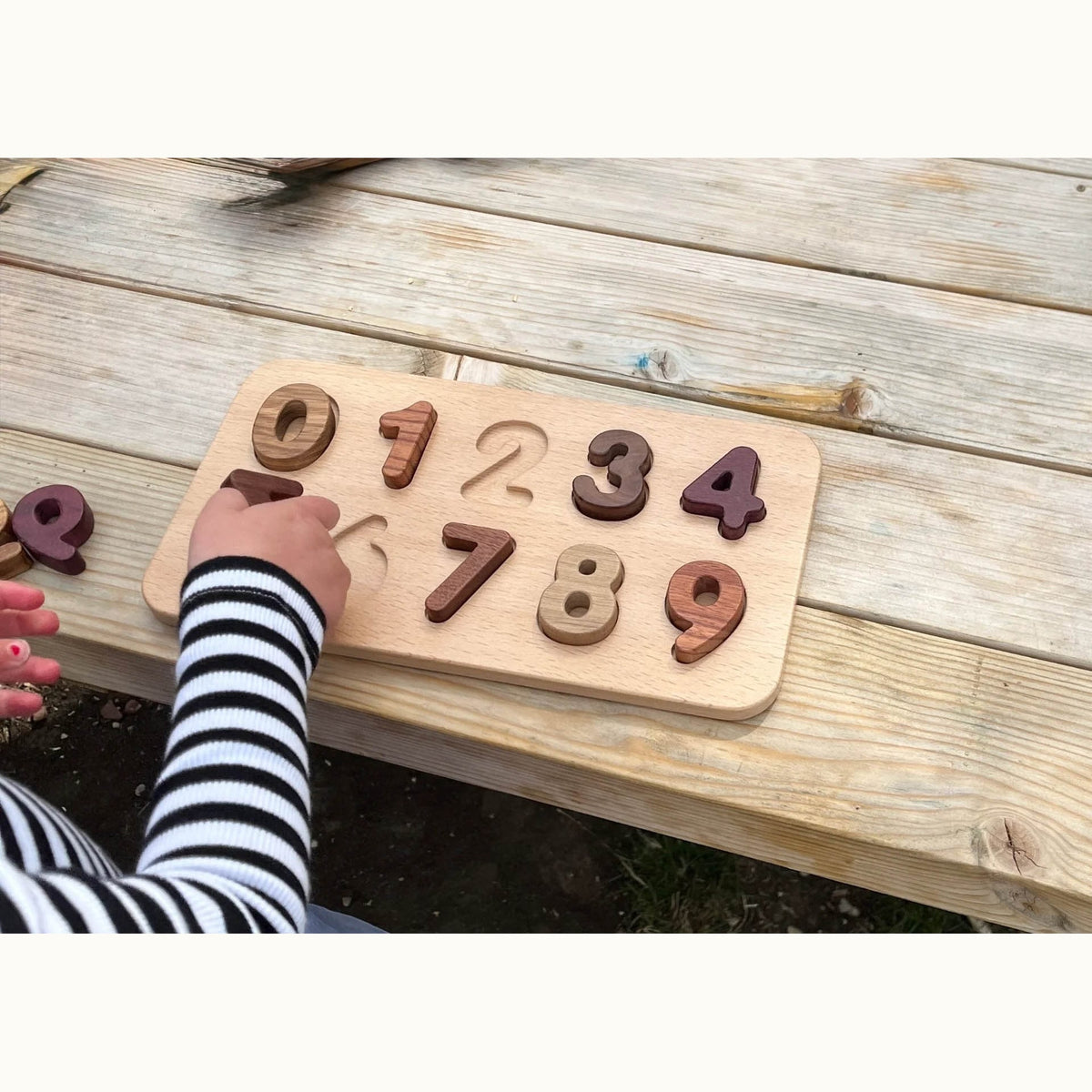 Child placing Walnut, Purpleheart & Tigerwood number puzzle pieces on beech board outdoors, digits 0–9 in natural hardwood to