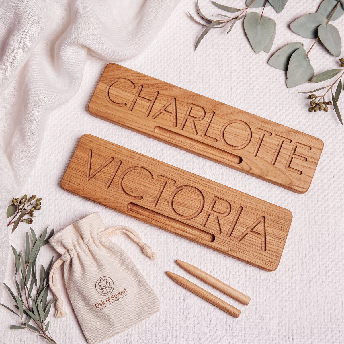 Two solid oak name tracing boards reading CHARLOTTE and VICTORIA, each with a stylus groove, beside two oak styluses and an Oak & Sprout cotton pouch