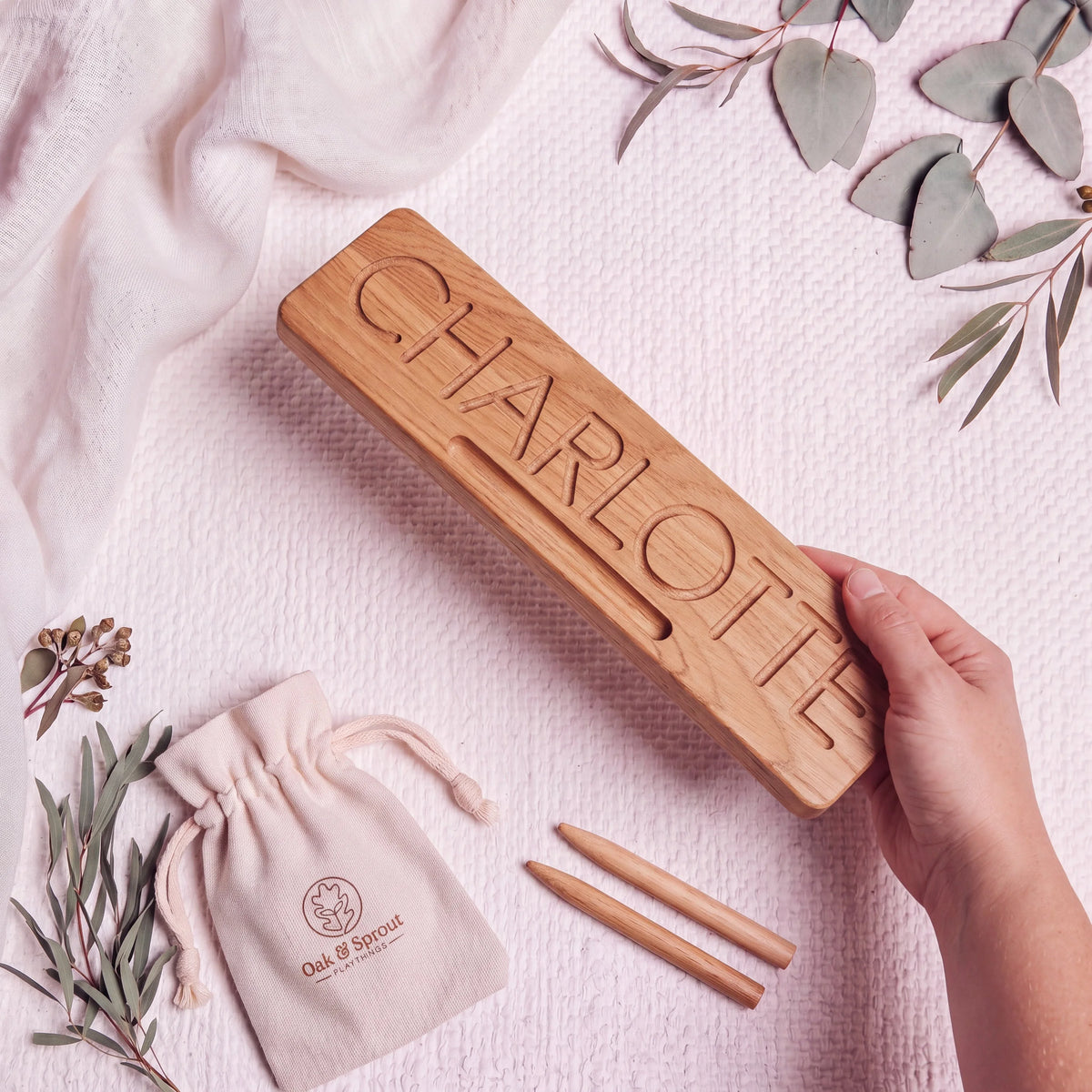 A hand holding a solid oak name tracing board reading CHARLOTTE, showing its thickness, with two oak styluses and an Oak & Sprout cotton pouch